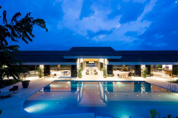 A luxurious poolside area at dusk, featuring an illuminated building with open seating, surrounded by sleek architecture and clear skies.