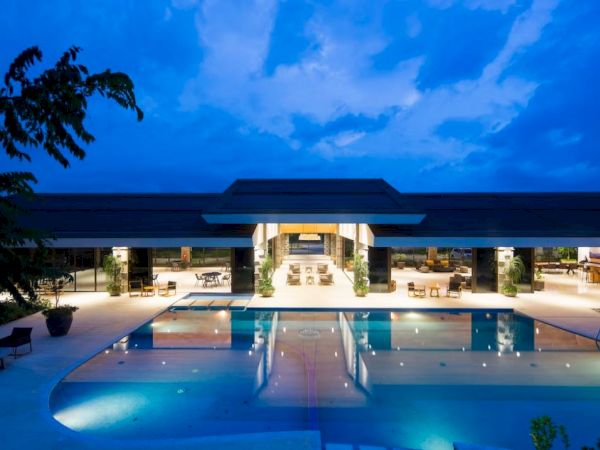 A luxurious poolside area at dusk, featuring an illuminated building with open seating, surrounded by sleek architecture and clear skies.