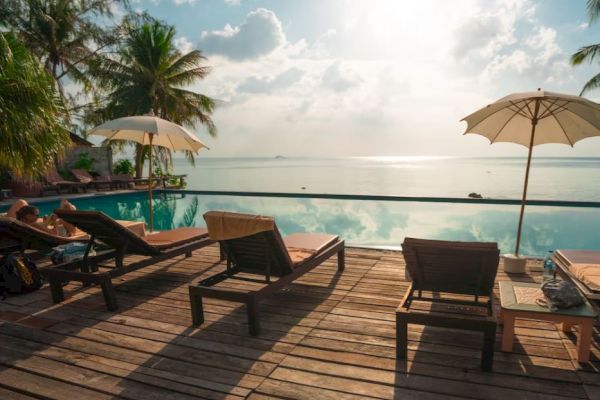 A serene poolside scene with lounge chairs, umbrellas, and palm trees overlooking an ocean view under a clear sky.