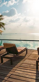 A serene beachside scene with wooden lounge chairs, umbrellas, a pool, and a scenic view of the ocean under a partly cloudy sky.