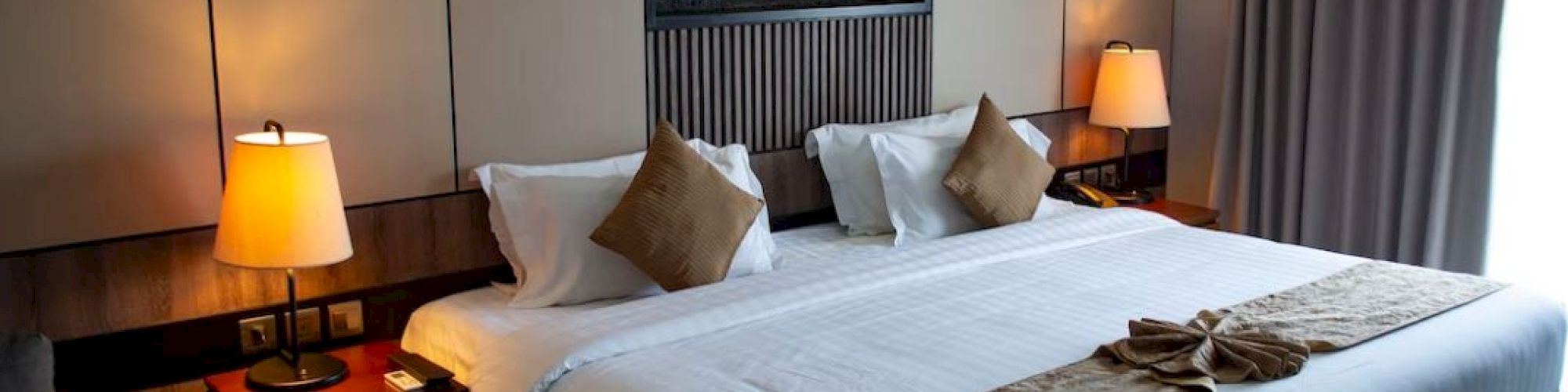 A well-lit hotel room with a large bed, side tables, lamps, and a wall-mounted decoration. Curtains cover a window on the right.