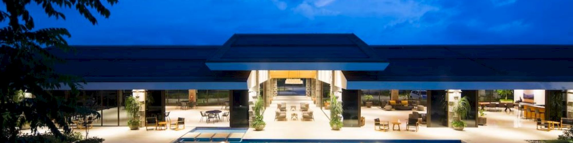 A modern building with large glass windows and a pool, set against a twilight sky.