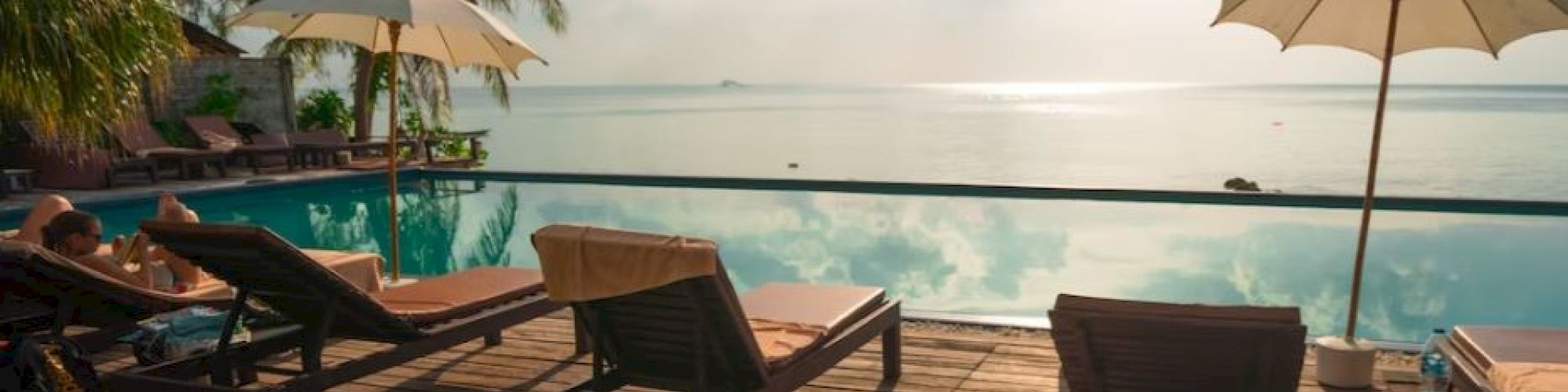 A serene poolside scene with sun loungers, umbrellas, palm trees, and a view of the ocean under a cloudy sky.
