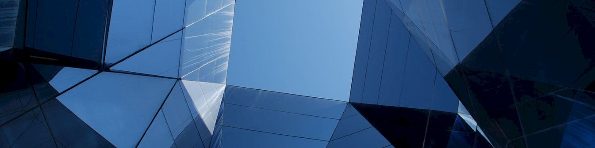A view looking up at geometric, reflective glass structures forming a symmetrical pattern against a clear blue sky.