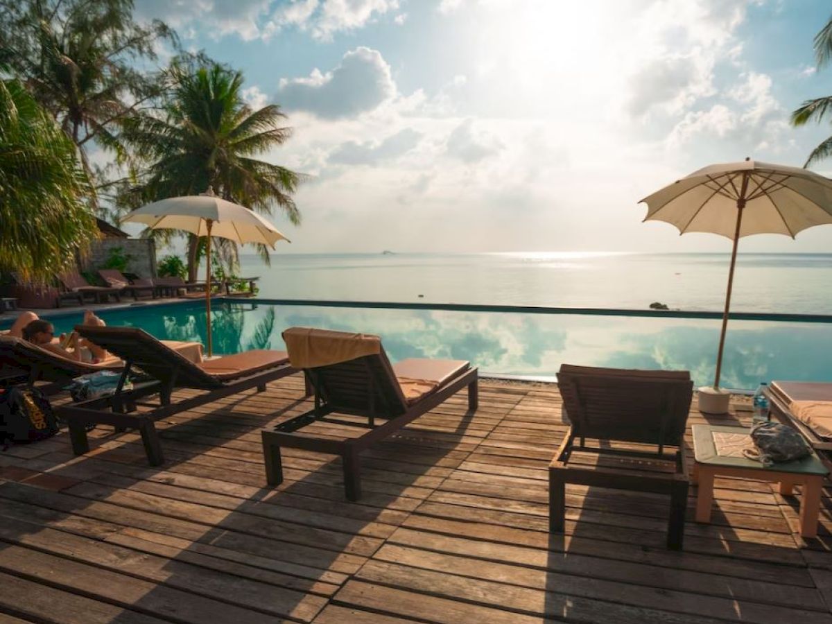A tropical beach scene with lounge chairs, umbrellas, palm trees, and a calm sea under a cloudy sky, viewed from a wooden deck.