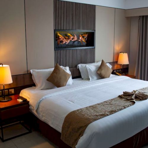 A modern hotel room with a king-sized bed, bedside tables, lamps, and a decorative wall feature. Curtains cover a window to the right.