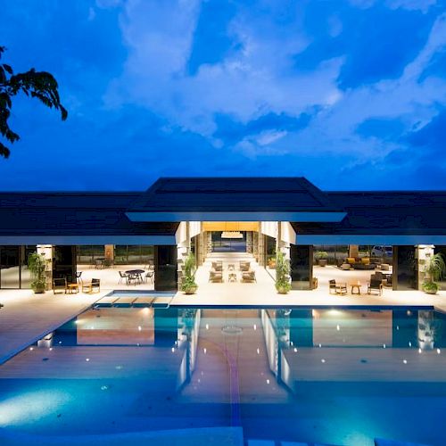 A modern building with large windows overlooks a well-lit pool area and lounge seating, set against a twilight sky.