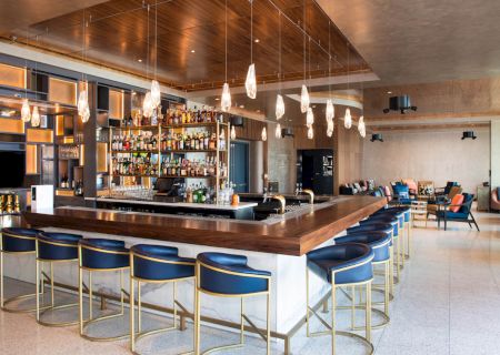 A modern bar with blue stools, hanging lights, and a well-stocked shelf. The room features wooden accents and a cozy seating area.