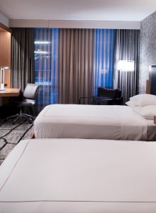 A hotel room with two beds, a large TV, desk, and modern decor, featuring neutral colors and ambient lighting in a spacious setting.