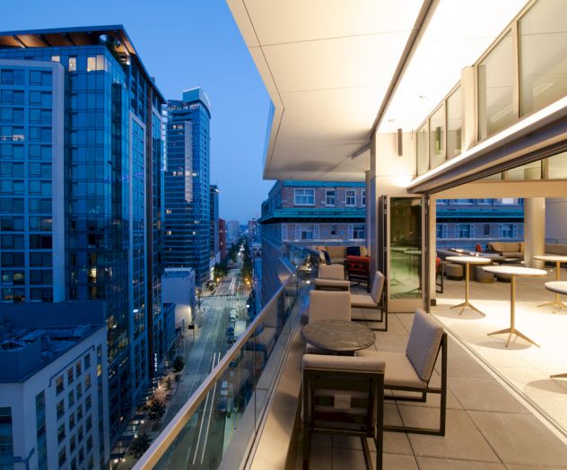A modern city balcony with tables and chairs overlooks a street lined with high-rise buildings at dusk.