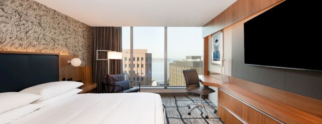 A modern hotel room with a large bed, TV, desk, and a window view of buildings and water is shown in the image.