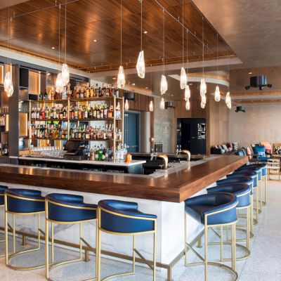 The image shows a modern bar with wooden accents, blue stools, a well-stocked liquor shelf, and pendant lights hanging from the ceiling.
