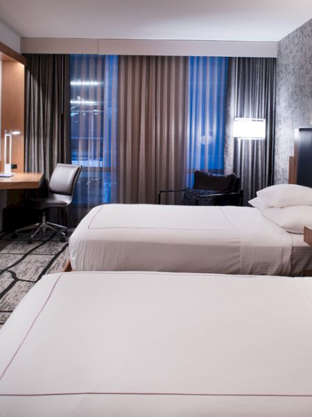 A modern hotel room with two beds, a flat-screen TV, desk, chair, and stylish decor. Warm lighting and patterned carpet complete the look.