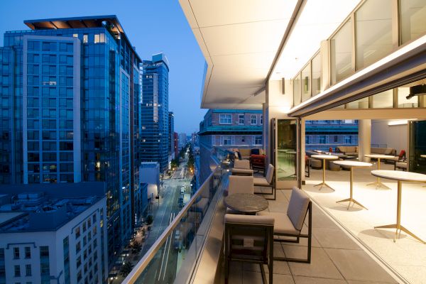 A modern balcony overlooks a city street surrounded by tall buildings at dusk, featuring seating areas with tables and chairs.