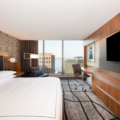 A modern hotel room with a large bed, desk, chair, and a TV. There's a window with a view of buildings and a body of water in the background.