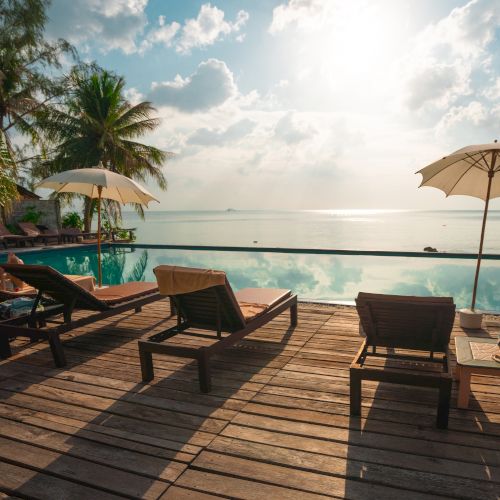 A serene beach scene with lounge chairs and umbrellas on a wooden deck by an infinity pool overlooking the ocean under a partly cloudy sky.