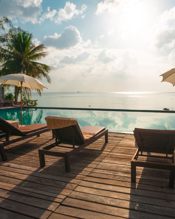 A serene beach scene with lounge chairs and umbrellas on a wooden deck by an infinity pool overlooking the ocean under a partly cloudy sky.