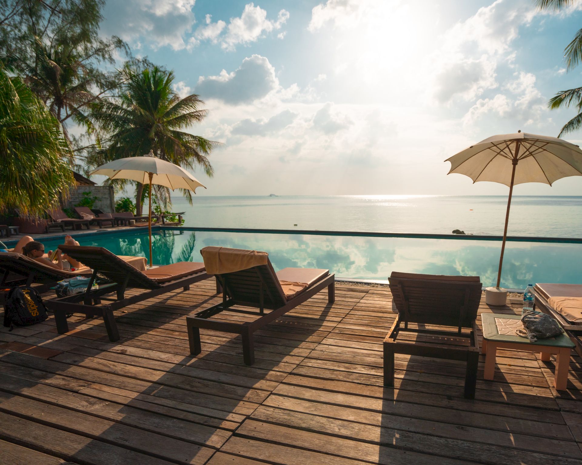 A serene beach setting with lounge chairs on a wooden deck, overlooking the ocean and framed by palm trees and umbrellas under a sunny sky.
