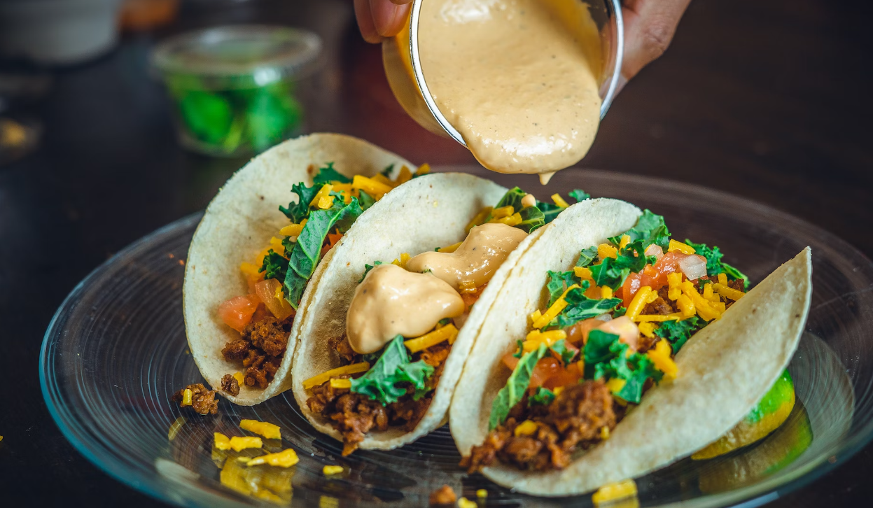 Three tacos filled with ground meat, cheese, kale, and tomatoes, being topped with creamy sauce on a clear plate.