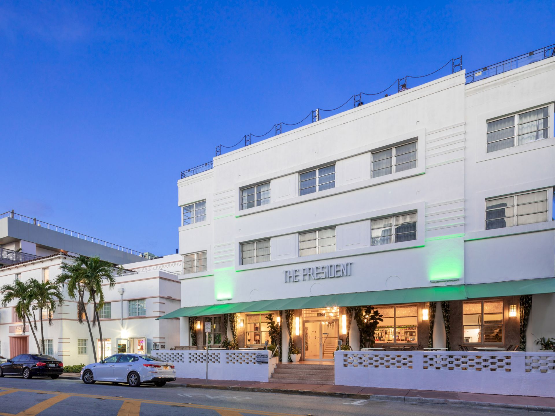 A white, multi-story building with a green awning and palm trees in front of it.