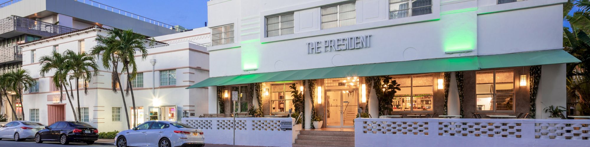 A modern white building, likely a hotel named "The President," with palm trees and parked cars, under a clear blue sky at dusk.