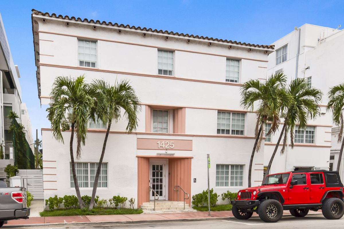 A white building with pink accents and a pink entrance, with palm trees and cars parked in front.