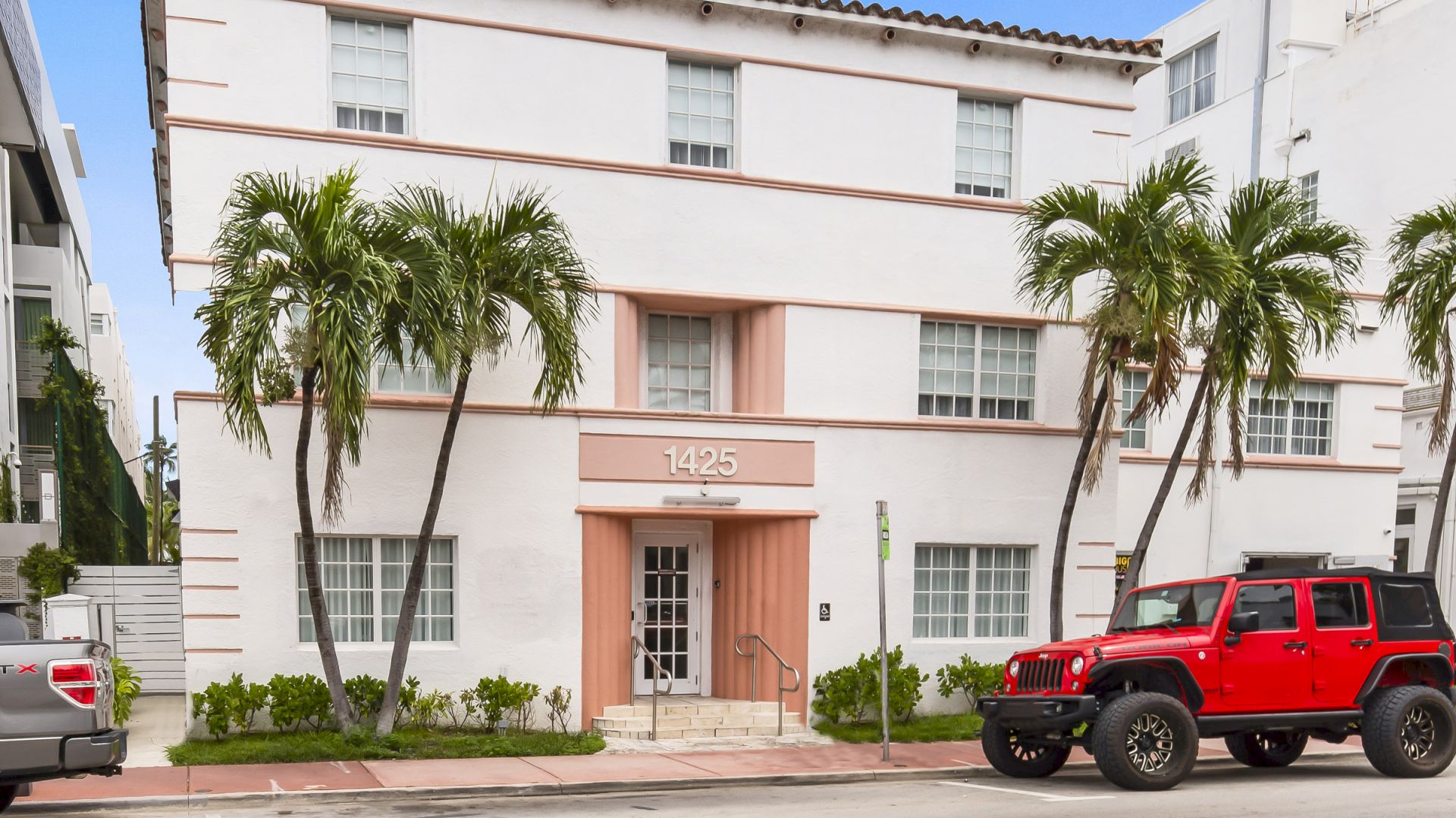 A three-story white building with pink accents, palm trees, and parked vehicles in front, including a red Jeep, under a blue sky.