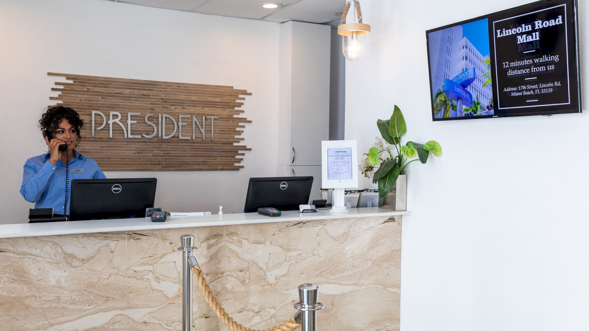A receptionist stands at a counter with two computers, a plant, and a wall-mounted screen showing "Lincoln Road Mall" information.