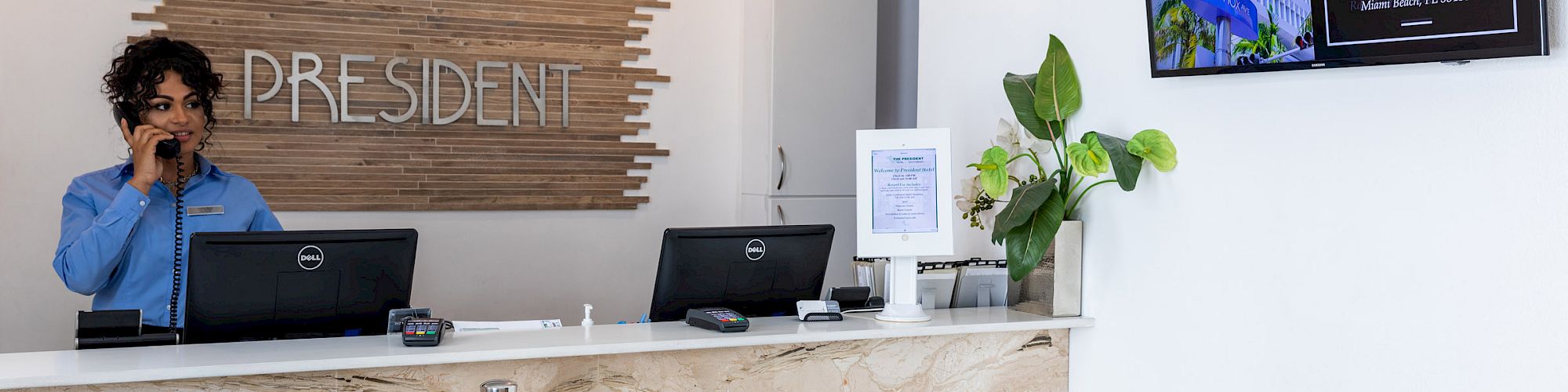 A receptionist stands at a counter with two computers, a plant, and a wall-mounted screen showing "Lincoln Road Mall" information.