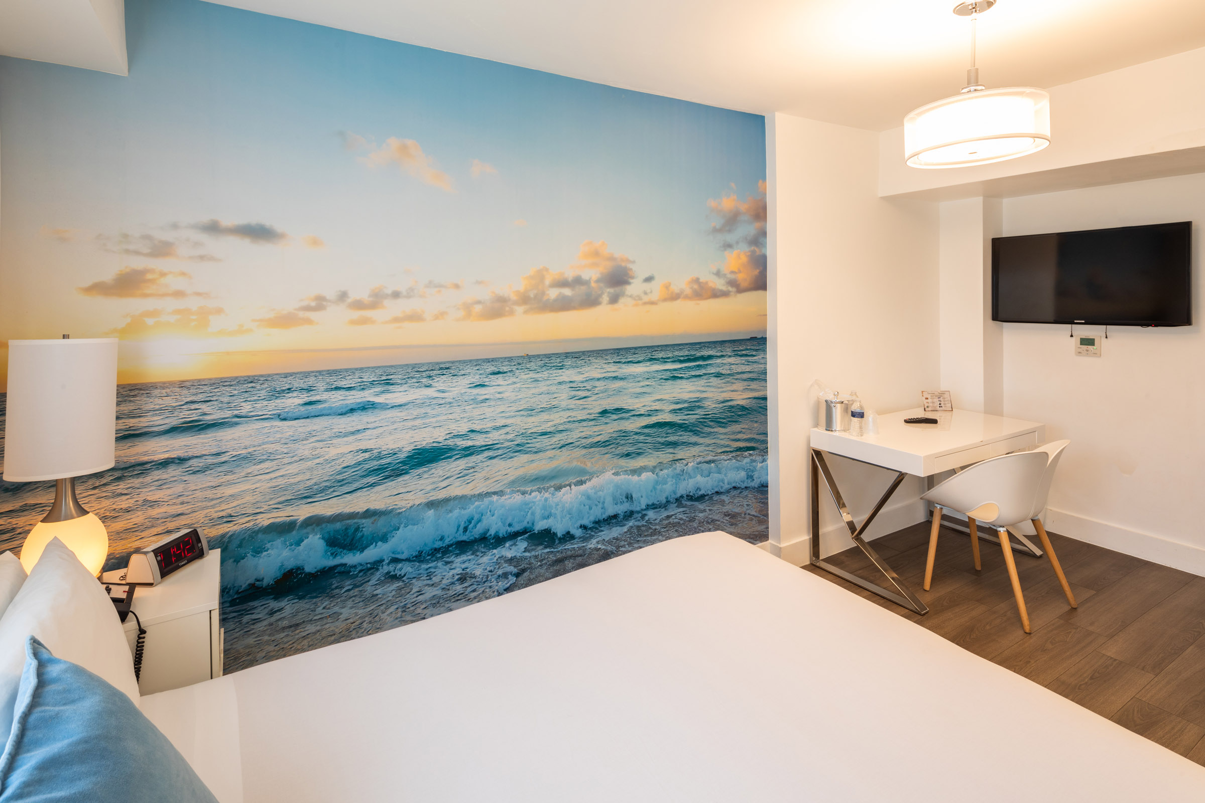 A hotel room with a large ocean view mural on the wall, a bed, desk, and TV