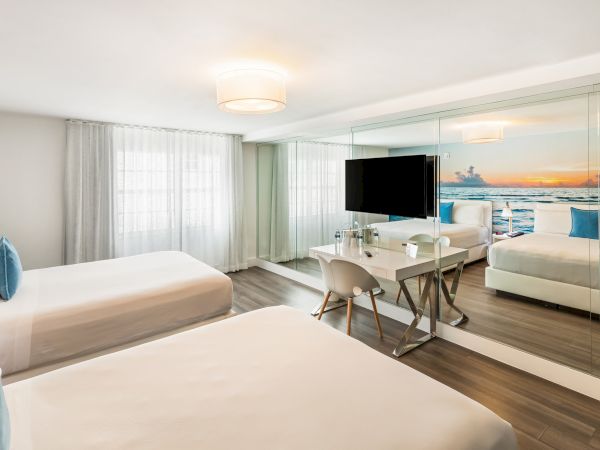 A modern hotel room with two beds, a large mirror, TV, and a scenic photo on the wall. The room is bright and stylishly decorated.