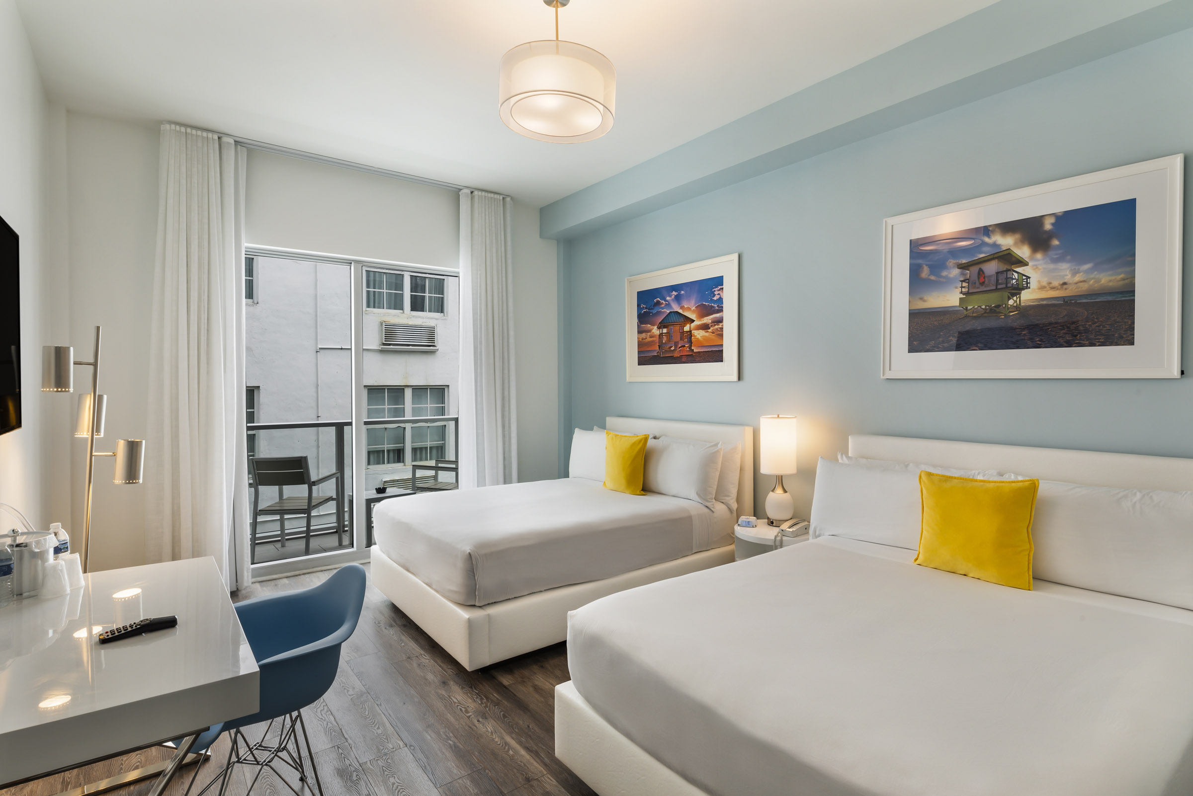 A hotel room with two beds, a desk, a balcony, and artwork on the walls.