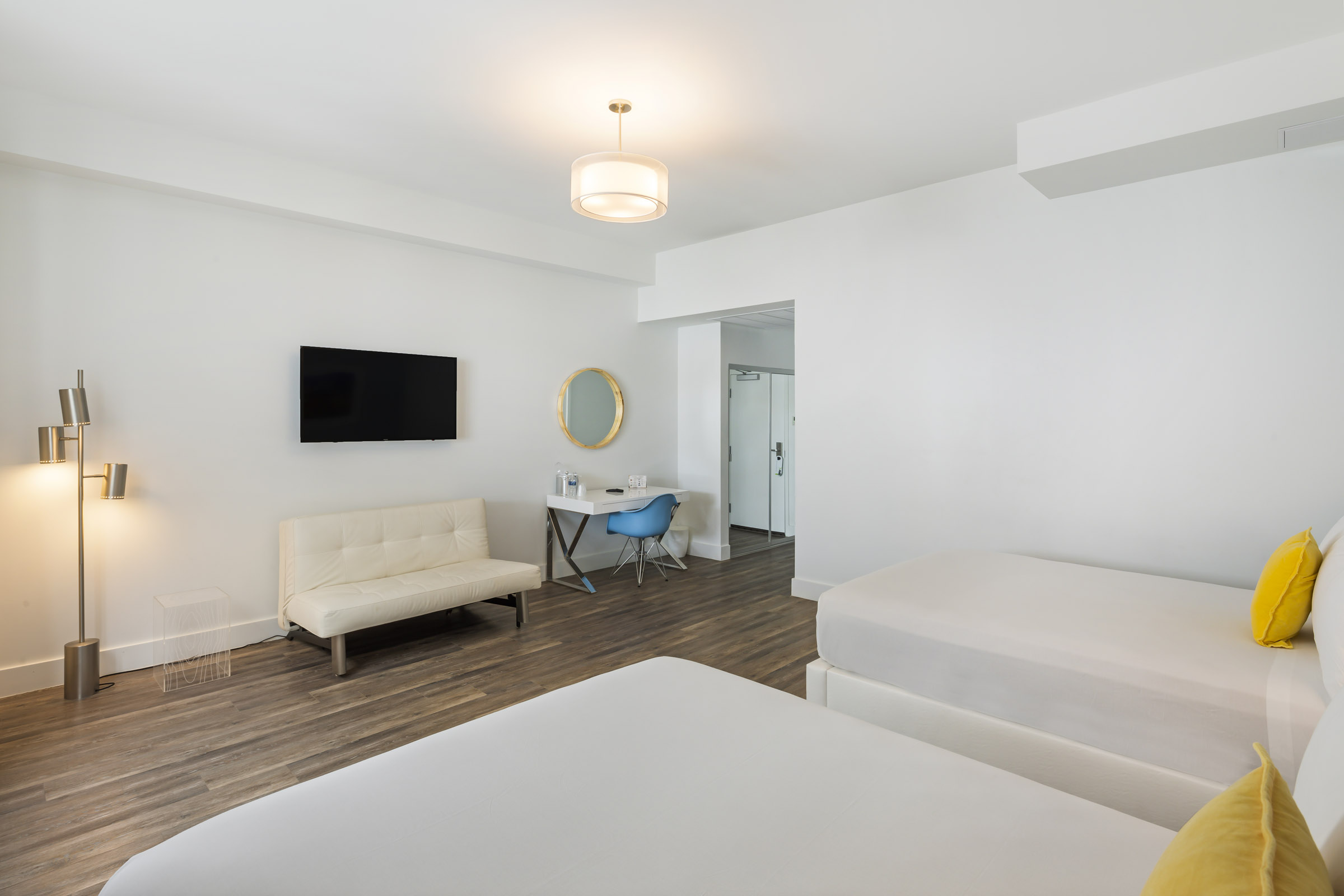 A clean and modern hotel room with two beds, a desk, a sofa, and a TV on the wall.