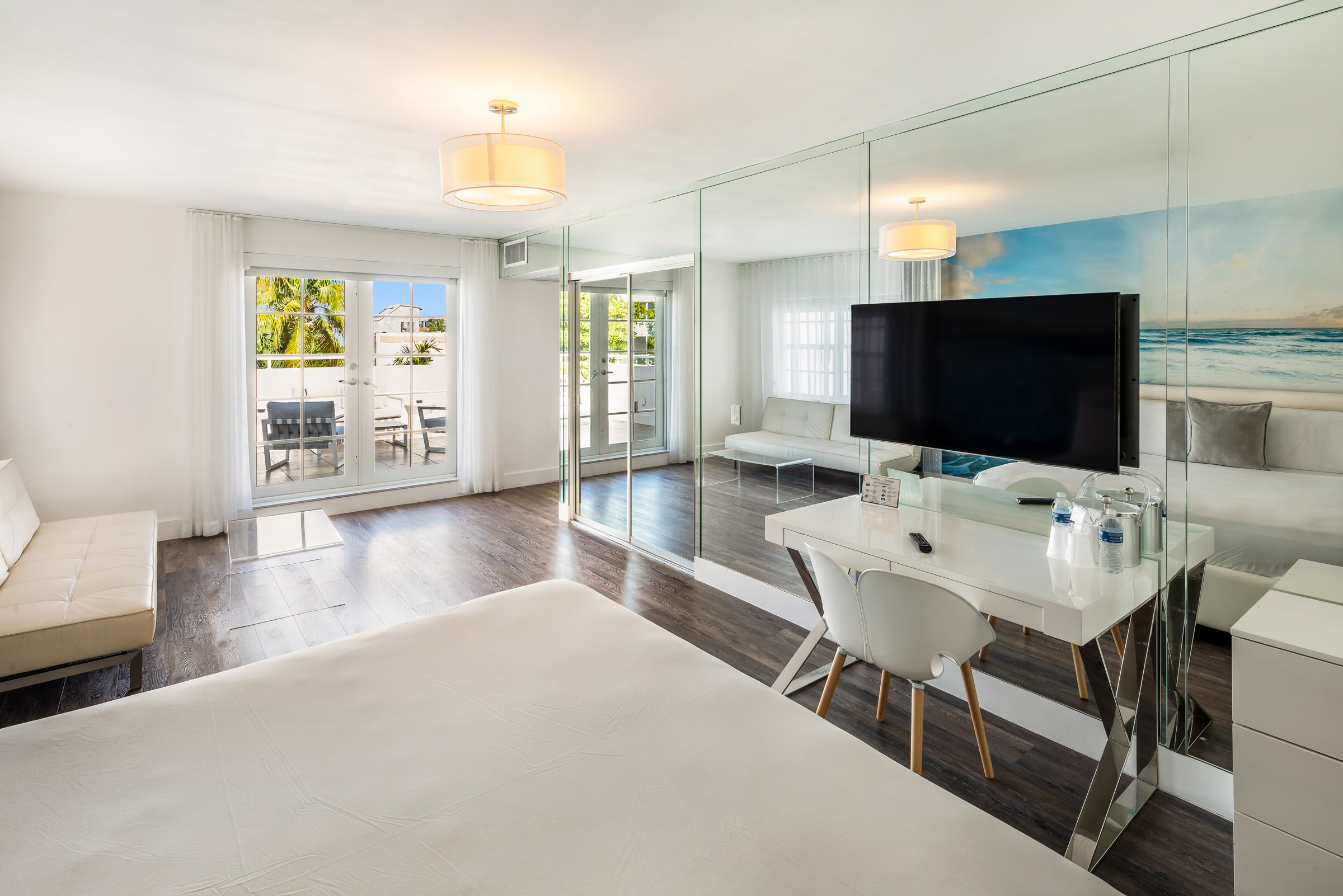 A modern hotel room with a large bed, desk, TV, and sofa, featuring a glass wall with a beach view and a balcony.