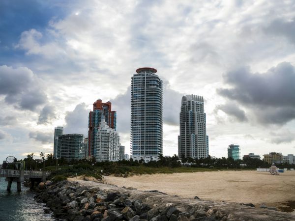 Tall buildings with a cloudy sky in the background, next to a rocky shoreline and sandy beach area.