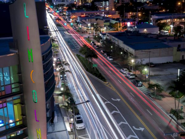 Cityscape at night featuring light trails from cars on a busy street, illuminated sign on a building, and high-rise buildings in the background.