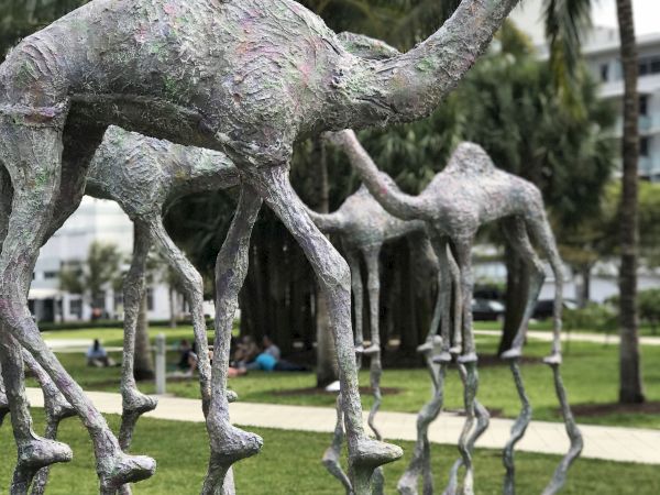 Sculptures of tall, abstract creatures with slender legs are displayed outdoors, set against a backdrop of palm trees and a grassy area.