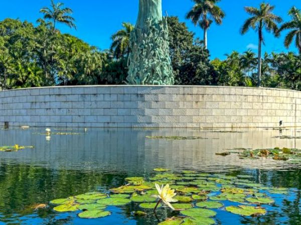 A sculpture rises from a circular stone platform surrounded by a water pond with lily pads, set against a backdrop of palm trees and clear blue sky.