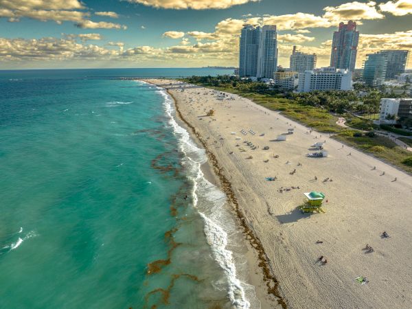 Aerial view of a beach with turquoise water, sandy shores, people, and tall buildings under a partly cloudy sky, possibly a coastal city.