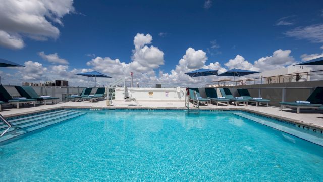 A large outdoor pool with lounge chairs and umbrellas under a blue sky with clouds.