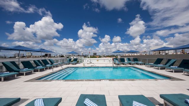 A rooftop pool with lounge chairs and umbrellas under a blue sky with clouds.