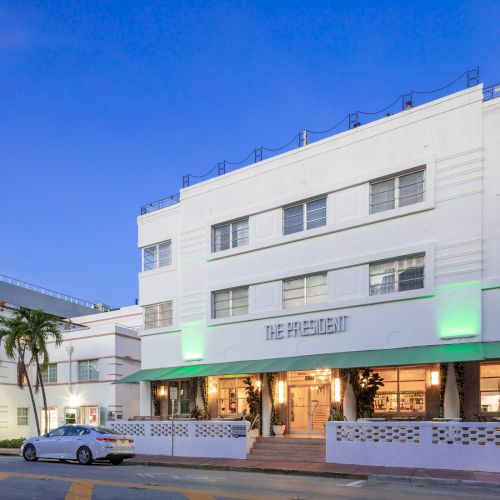 A white, multi-story Art Deco building labeled "The President" with cars parked outside, palm trees, and a soft evening sky in the background.