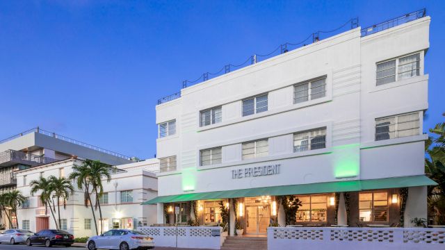 A white, multi-story building with a green awning and palm trees in front of it.