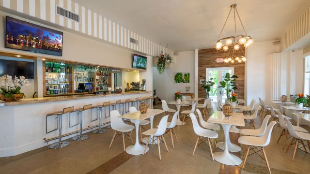 A brightly lit bar area with a counter, barstools, and a dining section with white chairs and tables.