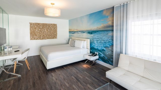 A wide view of a hotel room with a white bed, desk, sofa, and ocean-themed wall mural.