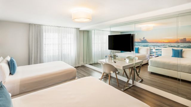 A wide view of a hotel room with two beds, a desk, and a mirror with a beach scene.