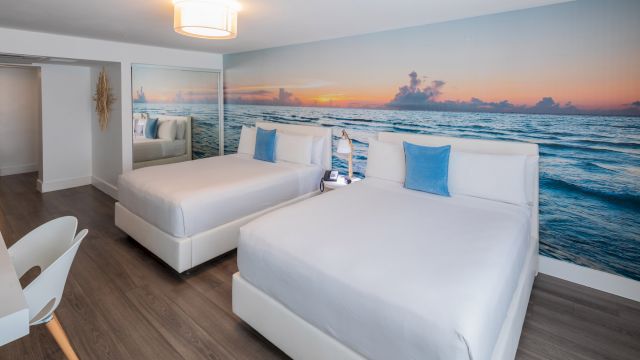 A clean and modern hotel room with two twin beds, a large wall mural of a beach scene, and a mirror.