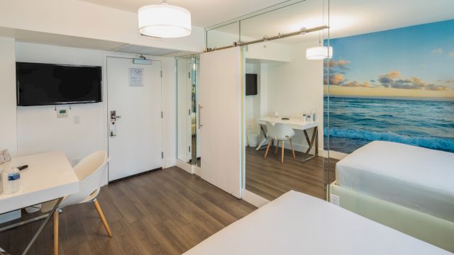 A hotel room with a large ocean view mural, a desk, chair, TV, and bed.