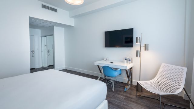 A white guest room with a bed, desk, chair, and TV.