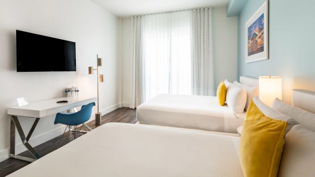 A hotel room with two beds, a desk, TV, and artwork.